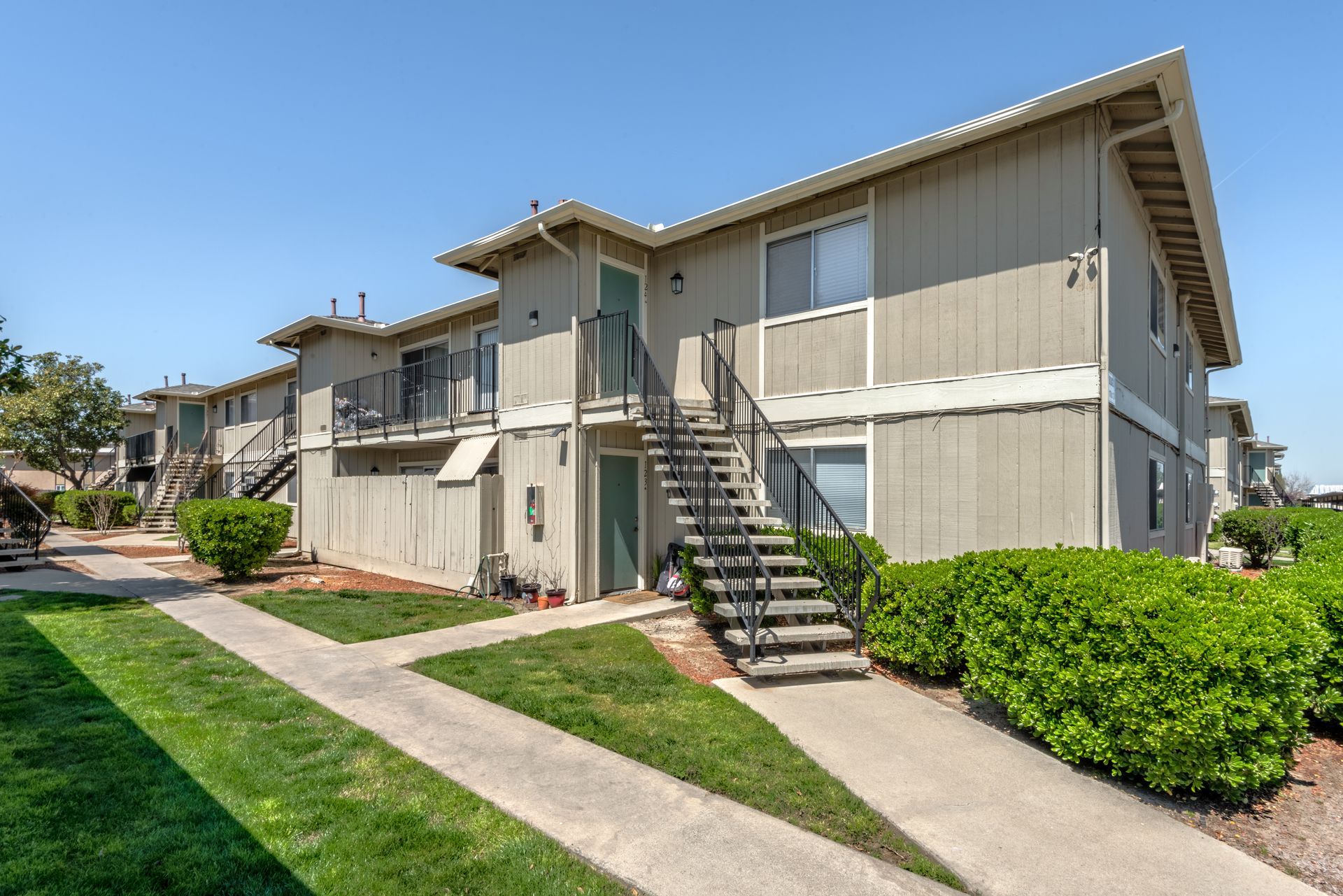 Apartment building exterior with stairs, tan siding, and green bushes. Sunny day.