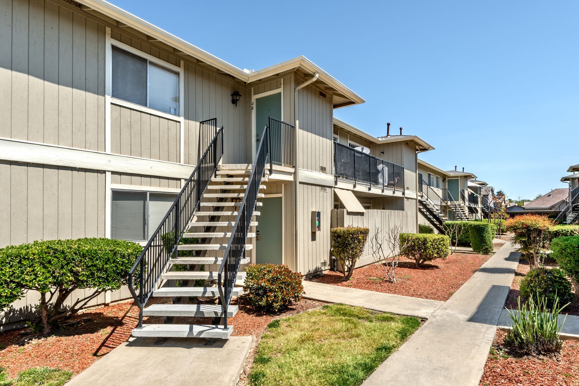 Two-story beige apartment building with outdoor stairways, green bushes, and a sunny day.