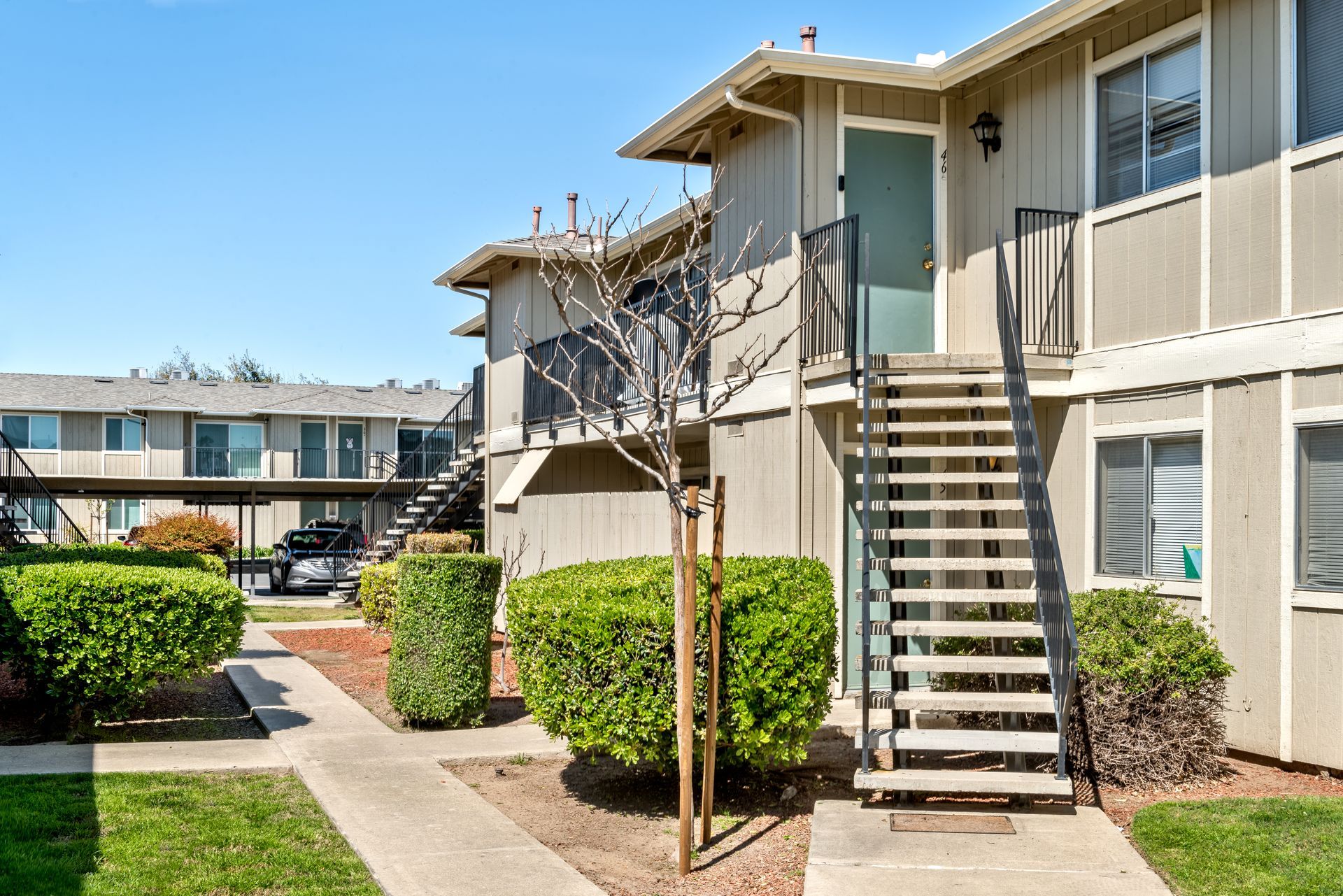 Apartment building exterior with stairs, walkways, and green landscaping under a clear blue sky.
