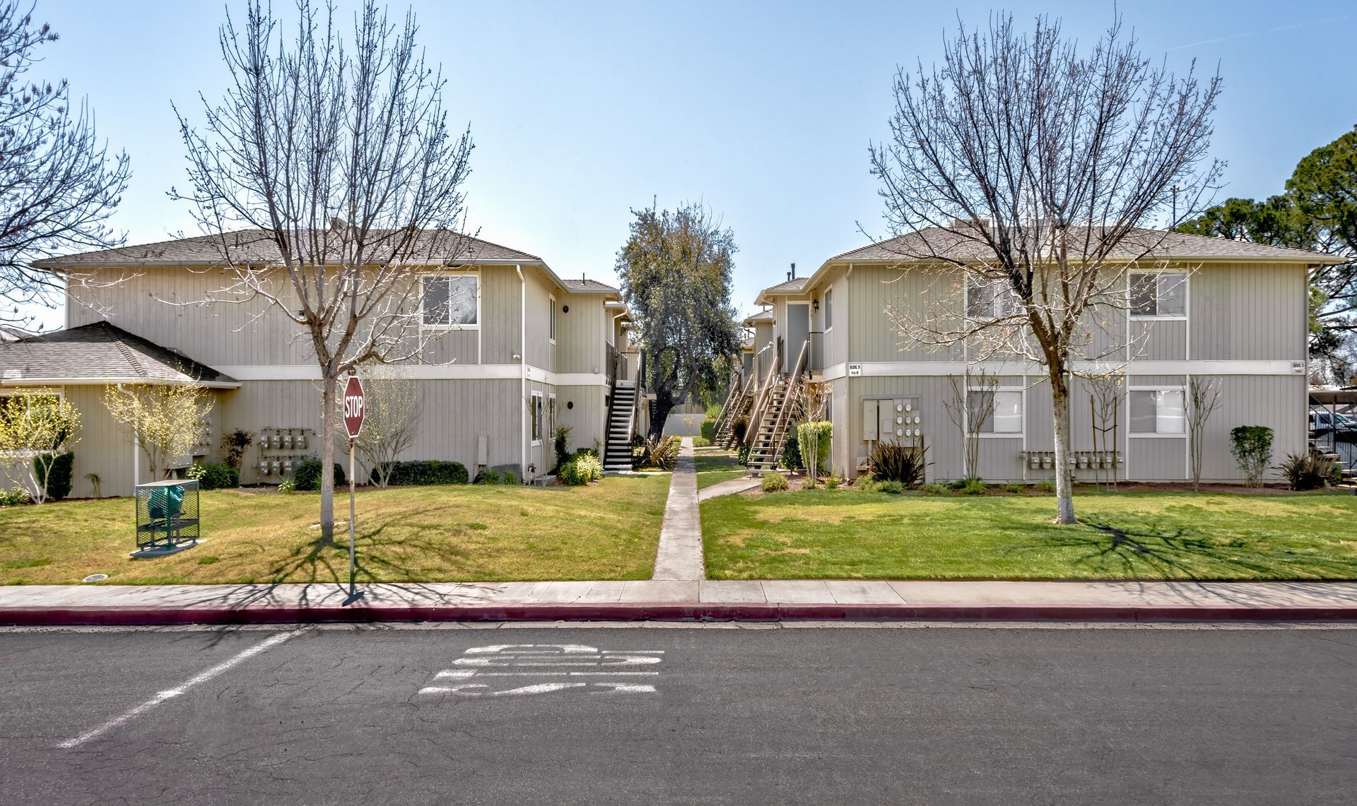 Two-story beige apartment buildings with green lawns and bare trees, viewed from the street.