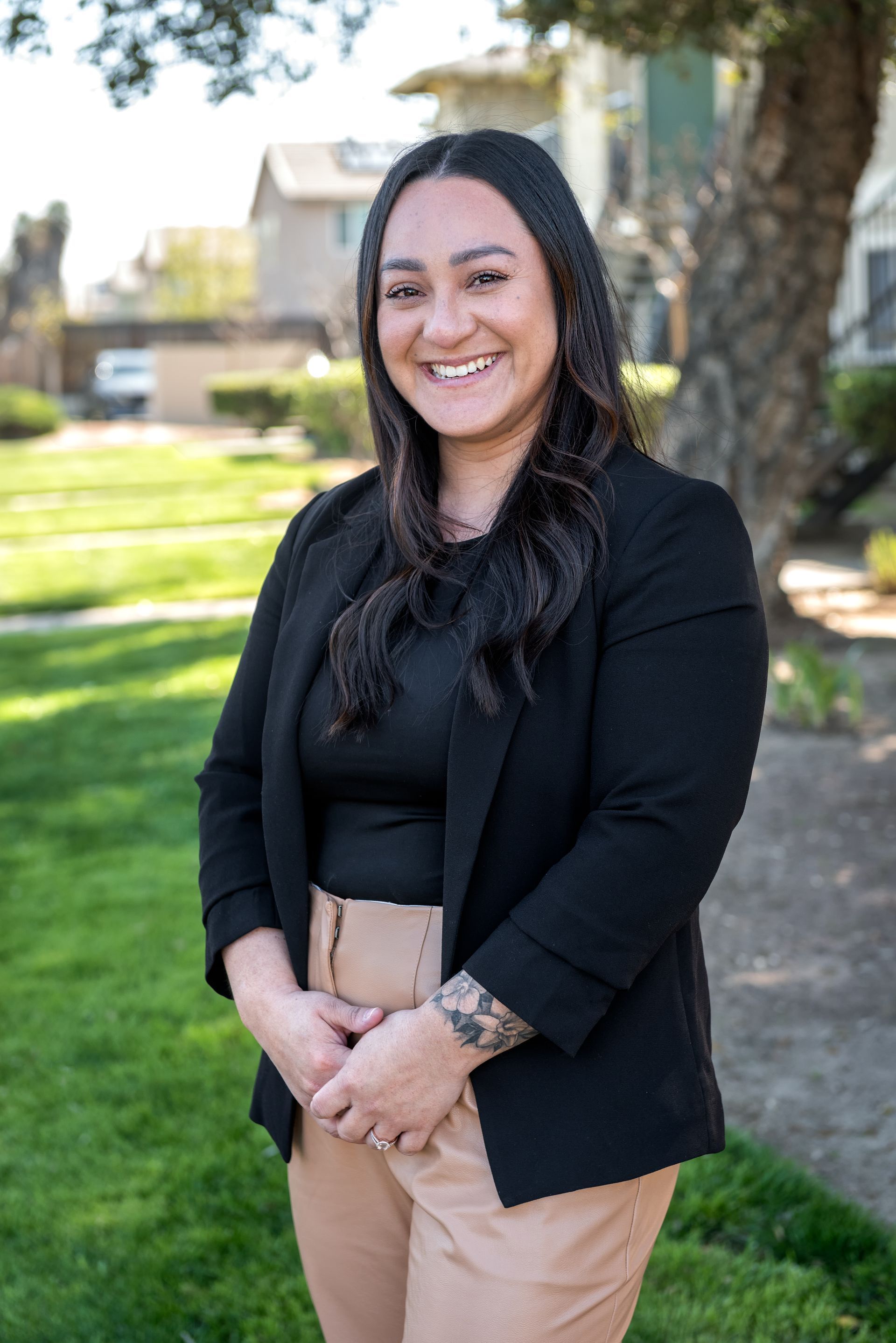 Woman with a tattoo smiles, wearing black blazer, tan pants, in front of grass and buildings.