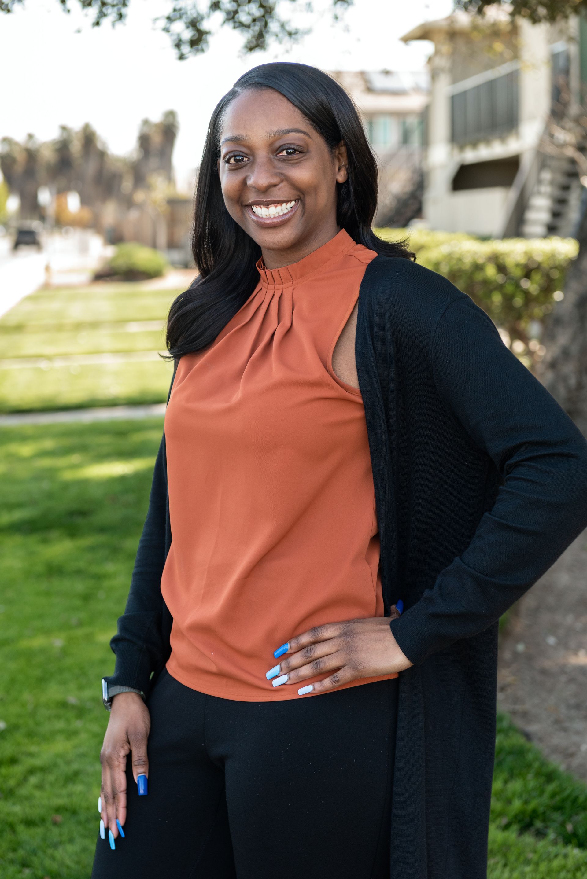 Woman in orange top and black cardigan smiles outdoors with hand on hip.