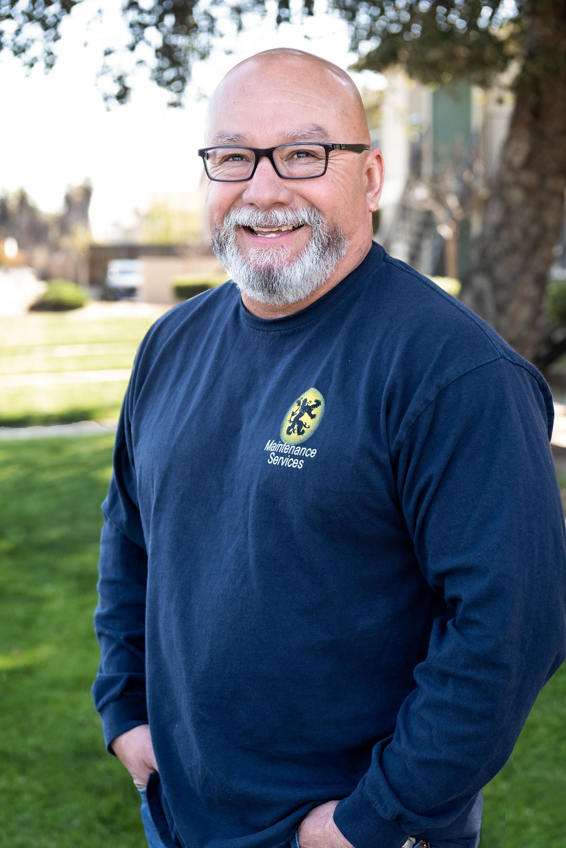 Mature man with glasses, gray beard, navy shirt, and hands in pockets smiles outdoors.