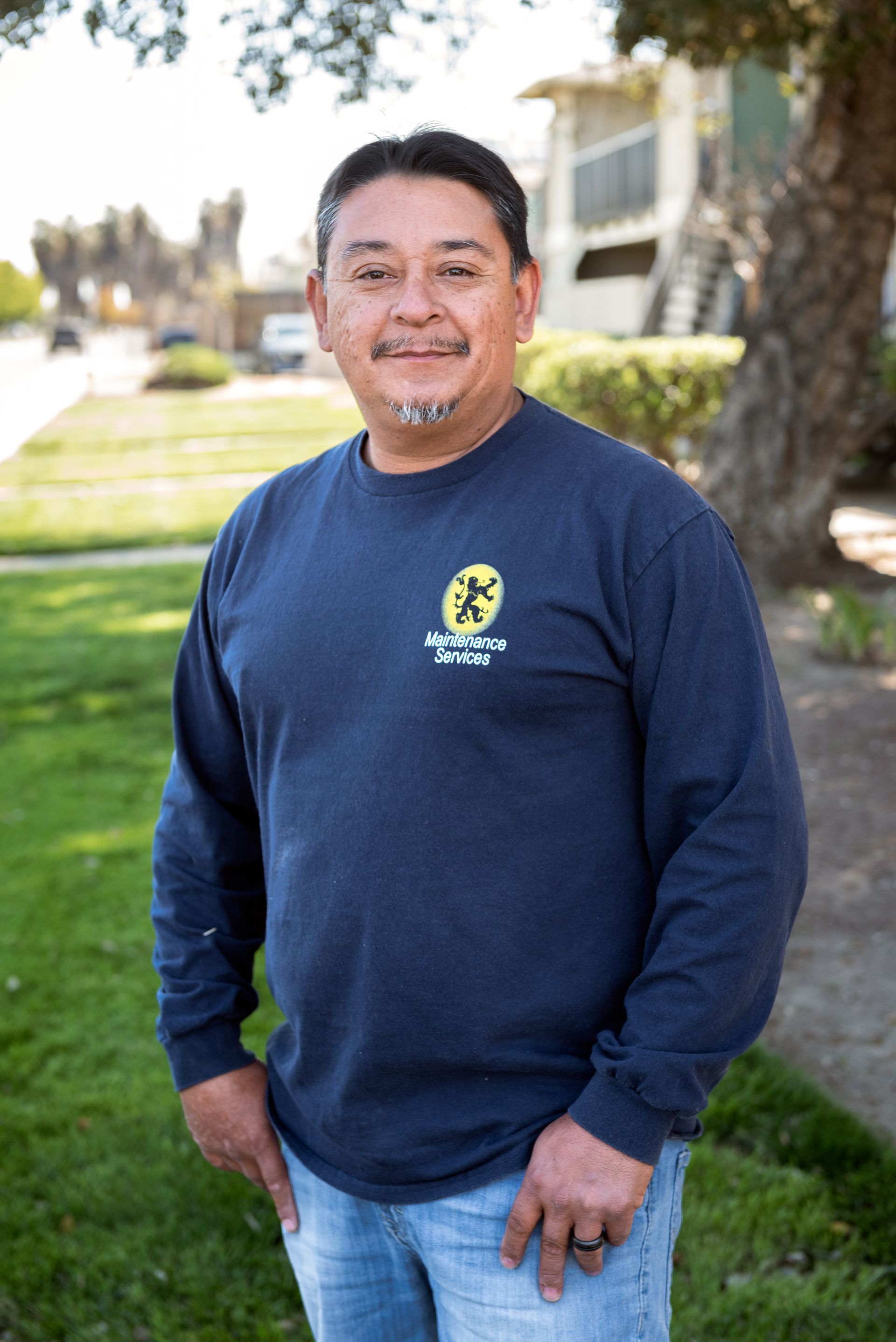 Man wearing a blue shirt and jeans, standing outside. He is smiling with hands in pockets.