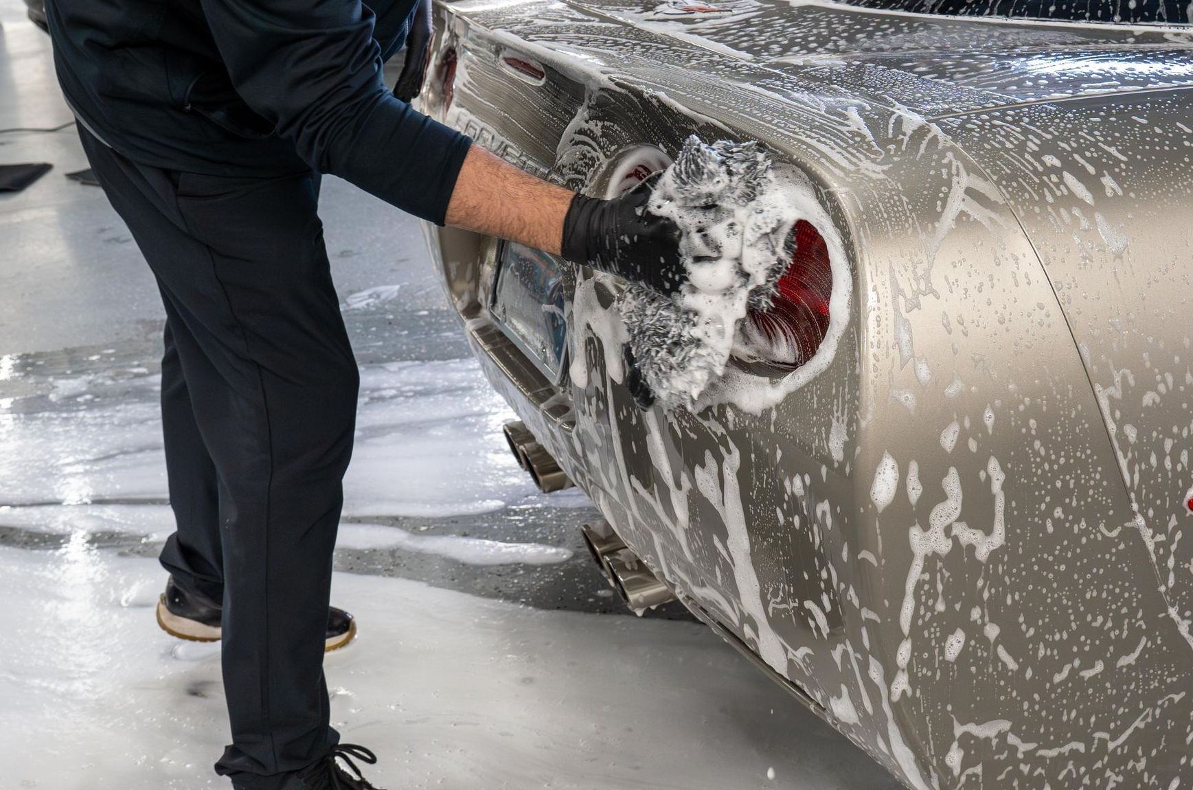 Person washing a silver car’s rear wheel with soapy water in a car wash