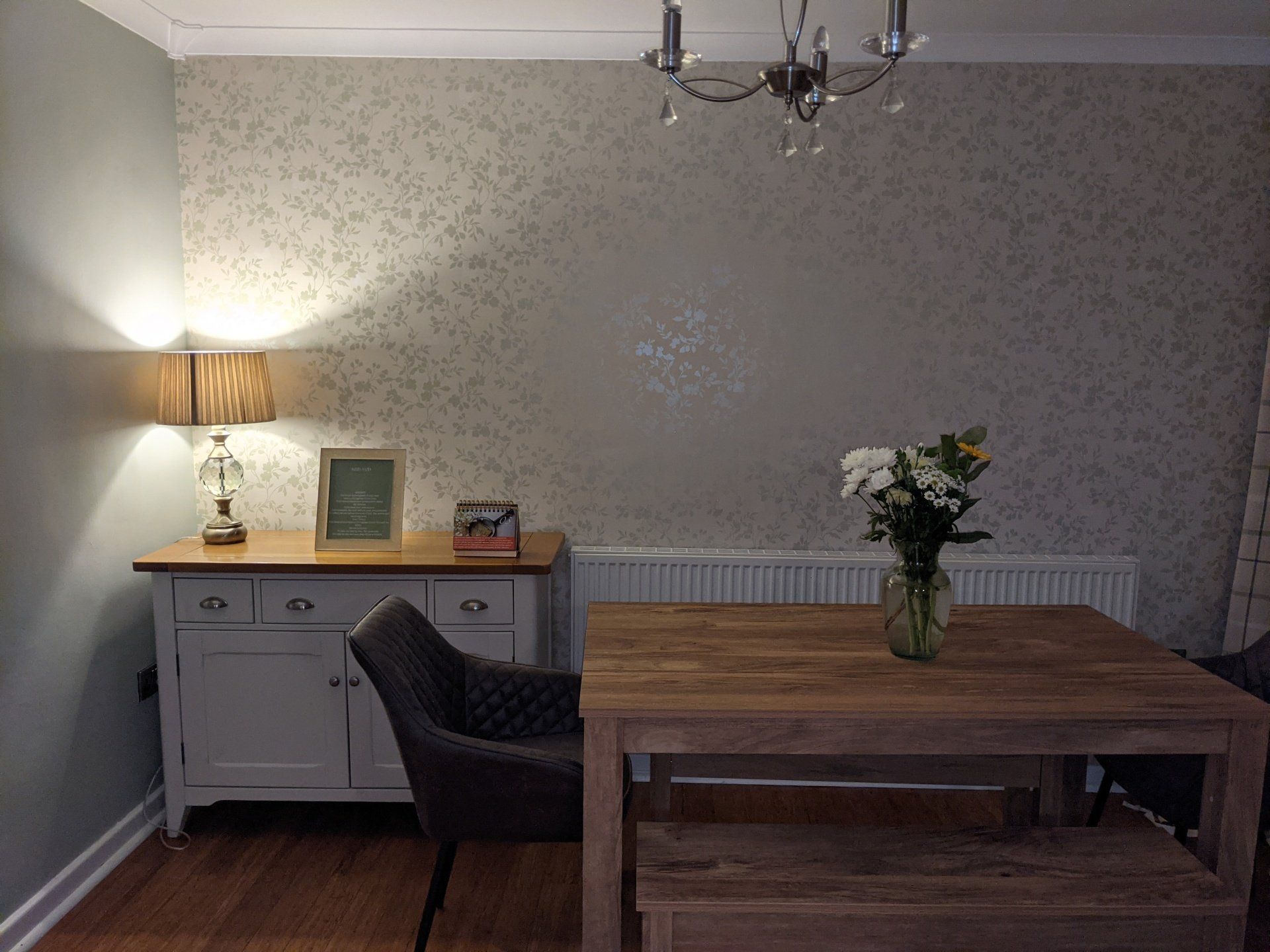 Picture of a modern kitchen diner painted in light green with floral designer wallpaper on the feature wall. There is a side table with lamp in the corner of the room.