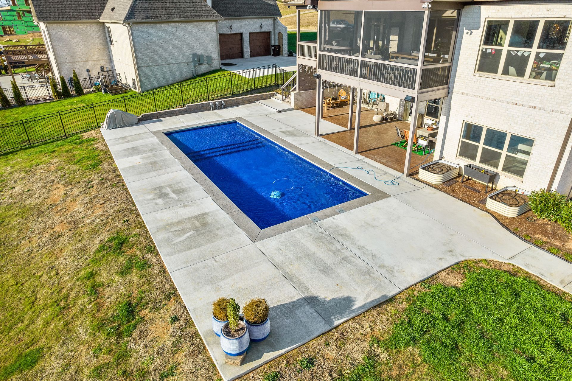 An elevated view of a rectangular pool surrounded by concrete patio near a two-story white house with a screened porch.
