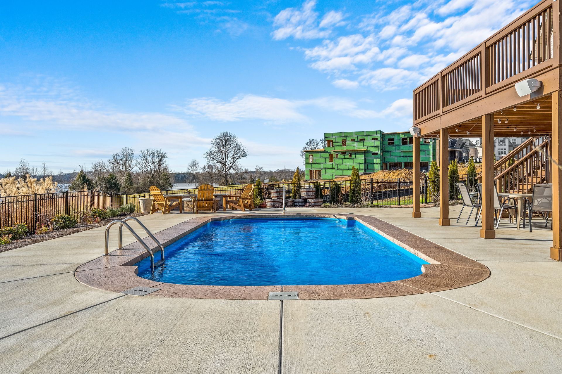 Pool in backyard with deck, trees, and lake view. Blue water, sunny sky.