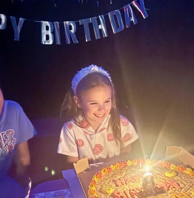 A little girl is sitting in front of a birthday cake under a happy birthday banner
