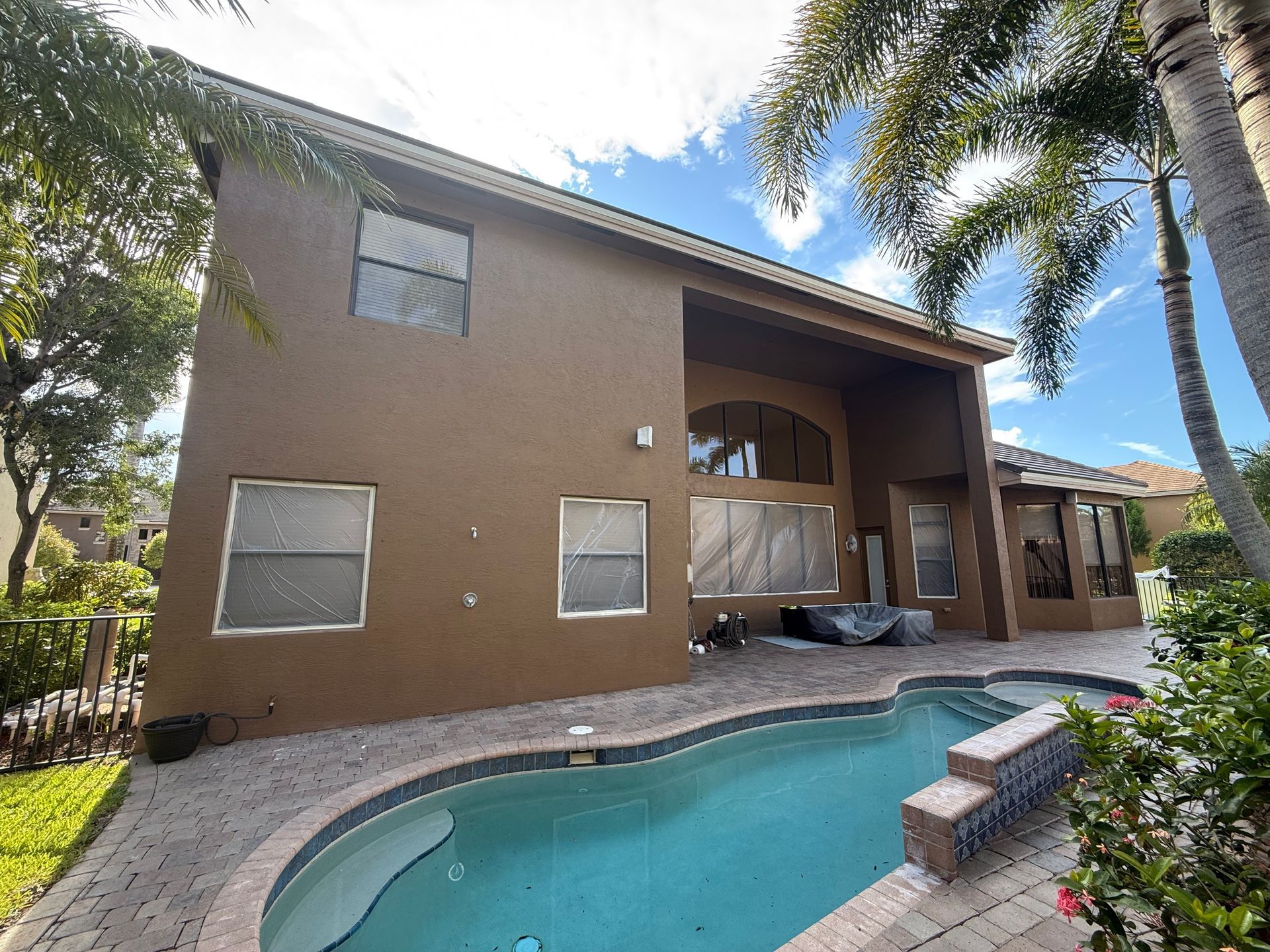 Two-story brown house with a pool and palm trees under a blue sky.