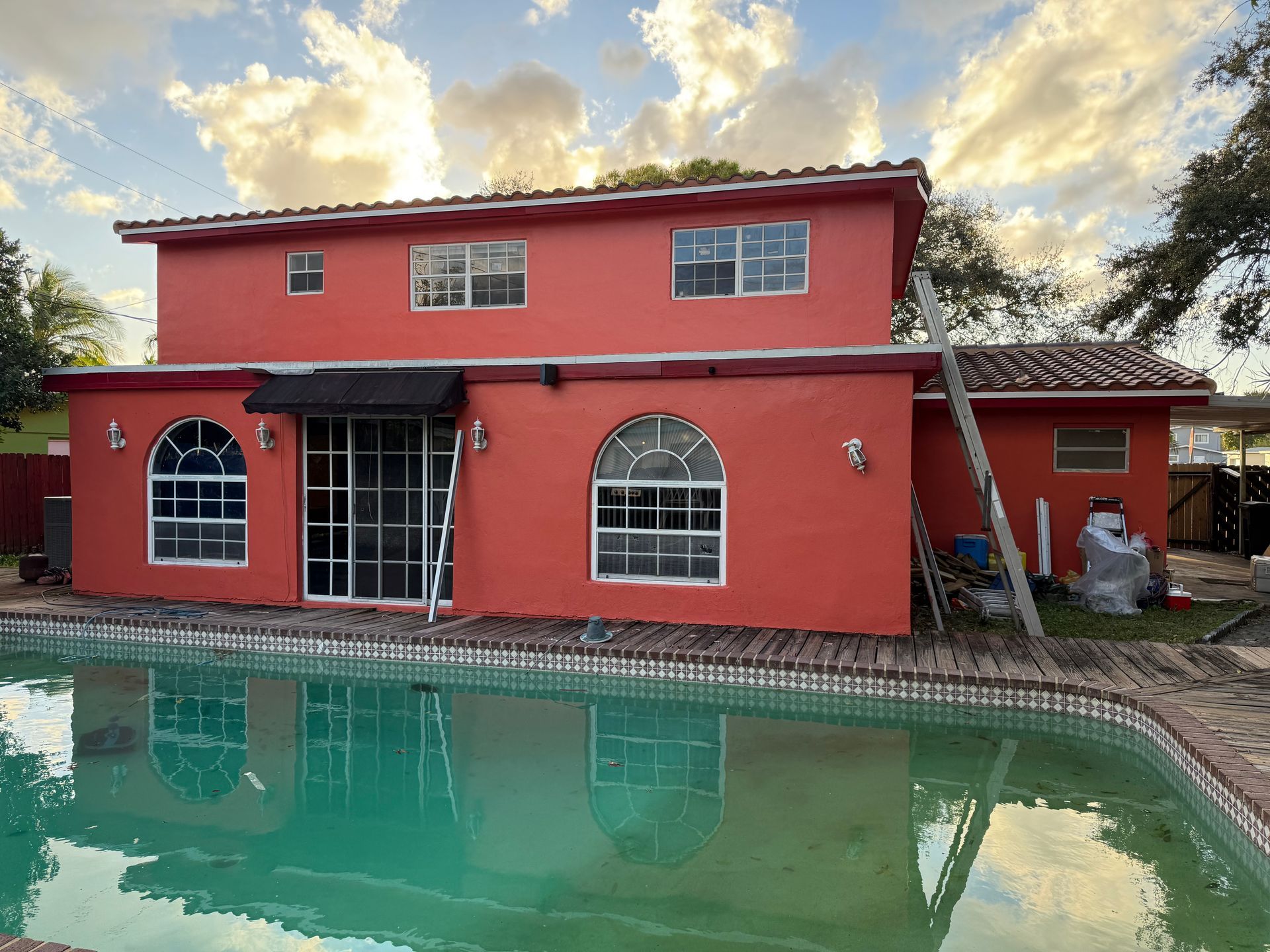 Red two-story house with a pool. Windows and awning visible. Reflective pool water with cloudy sky background.