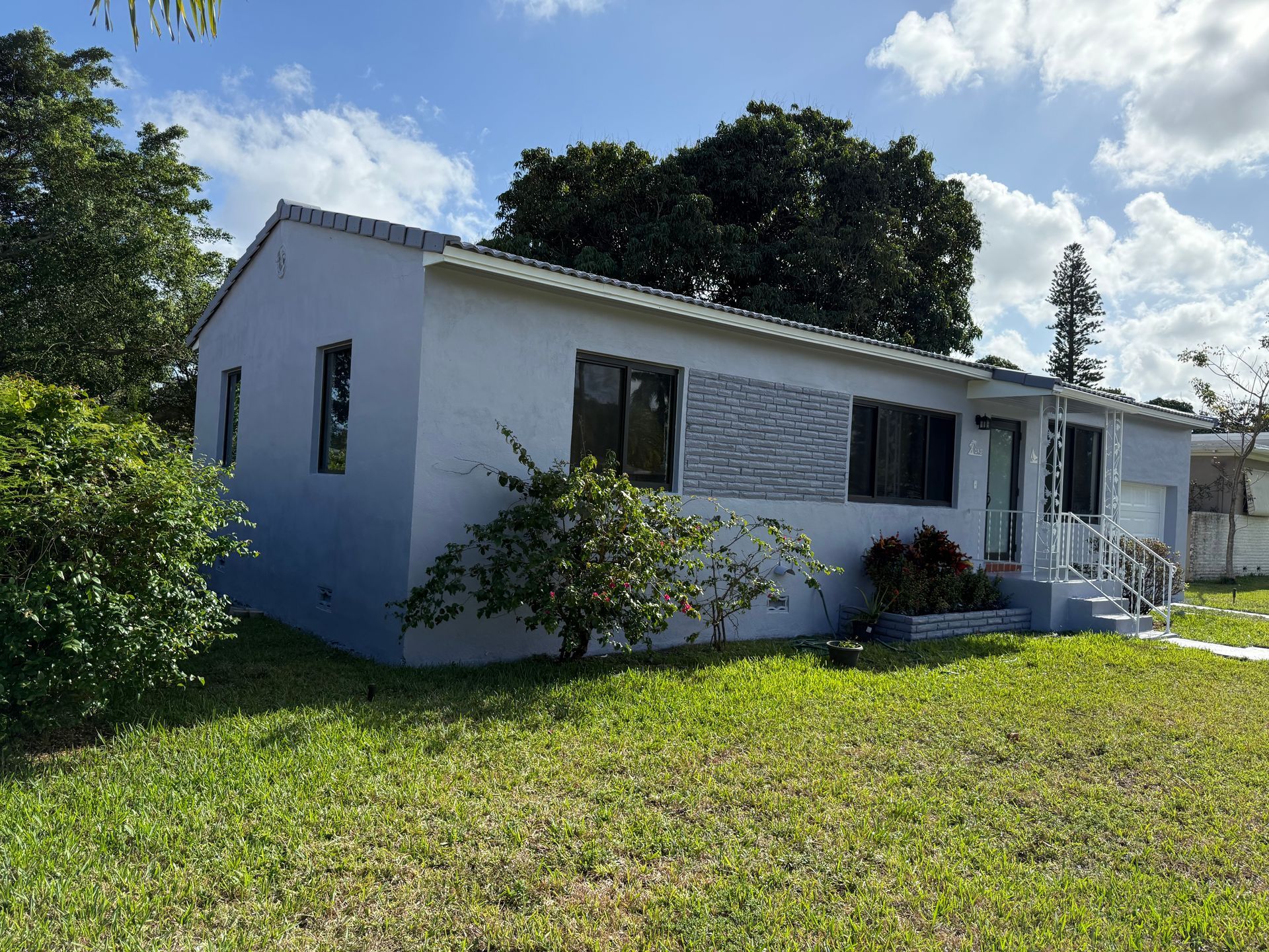 Gray house with windows, front door, and a small porch on a grassy lot with trees and bushes.
