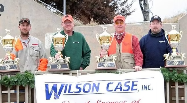 Four men holding trophies stand behind a banner that reads