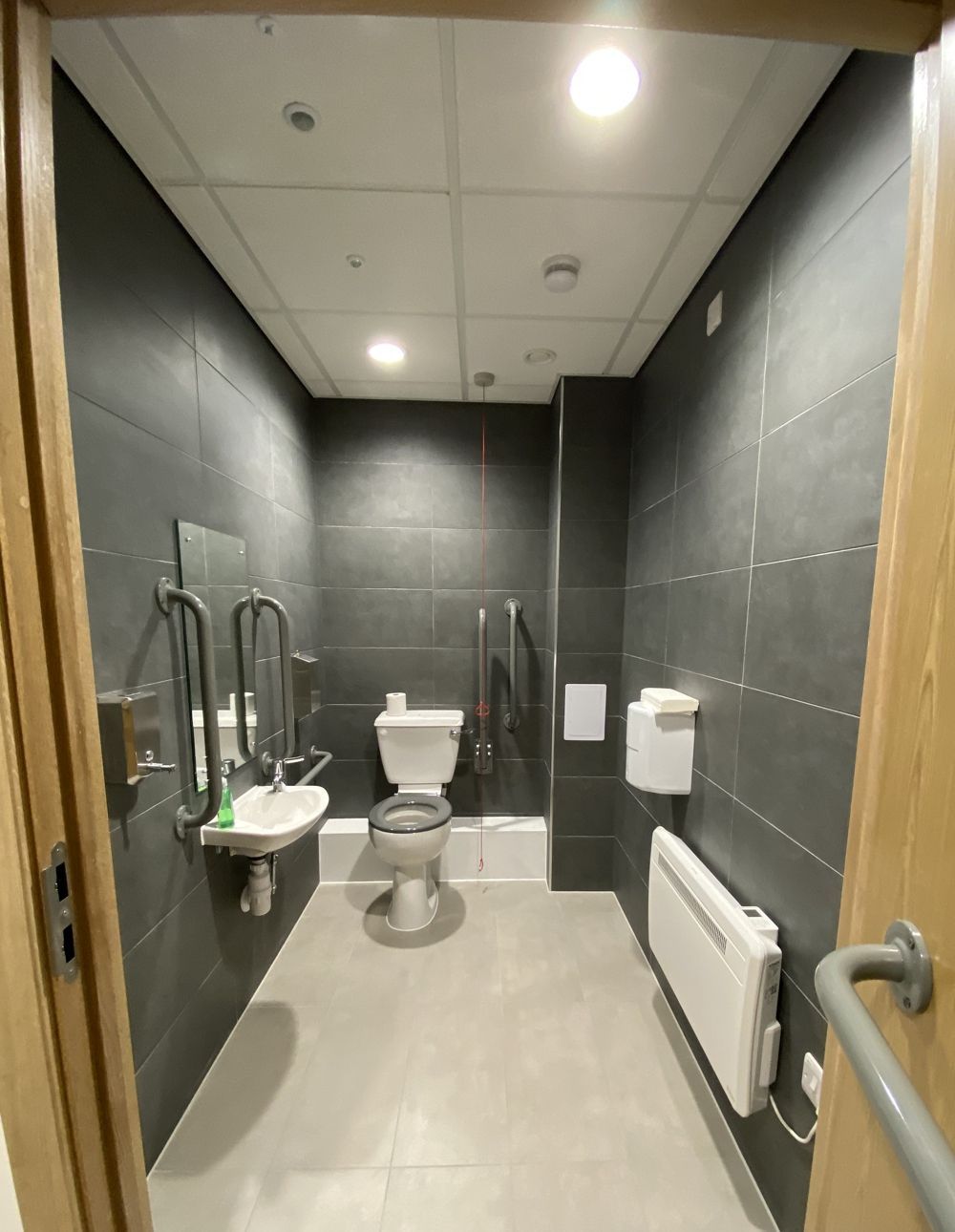 Accessible public restroom with gray tiled walls, toilet, sink, and grab bars.