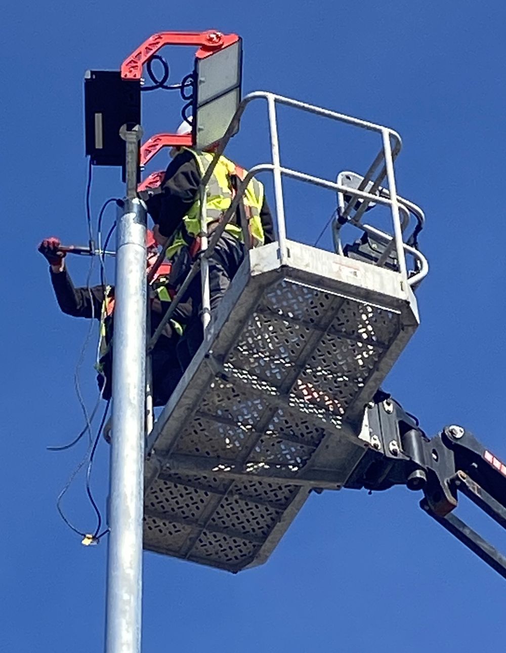 A worker on a lift installs equipment on a pole. Wearing a safety vest, against a blue sky.