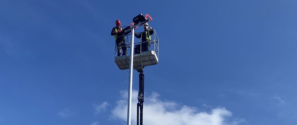 Two workers in a lift bucket installing equipment on a pole against a blue sky.