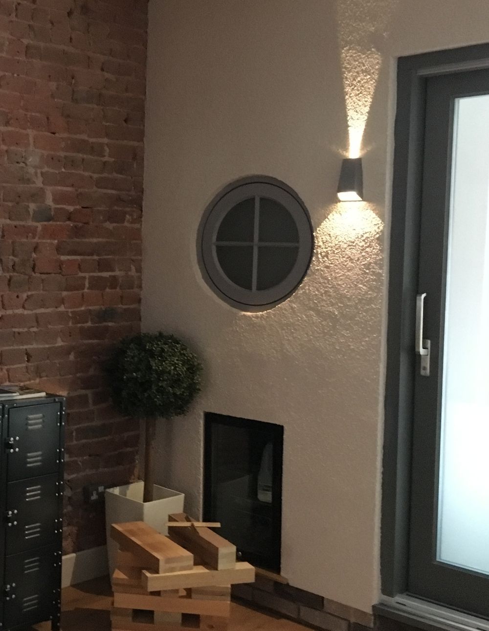 Interior corner with brick wall, round window, up-lighting, a door, and a decorative topiary.