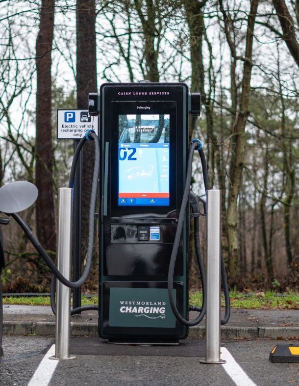 Electric vehicle charging station with touchscreen display, cords, and metal poles; outdoors in front of a parking spot.