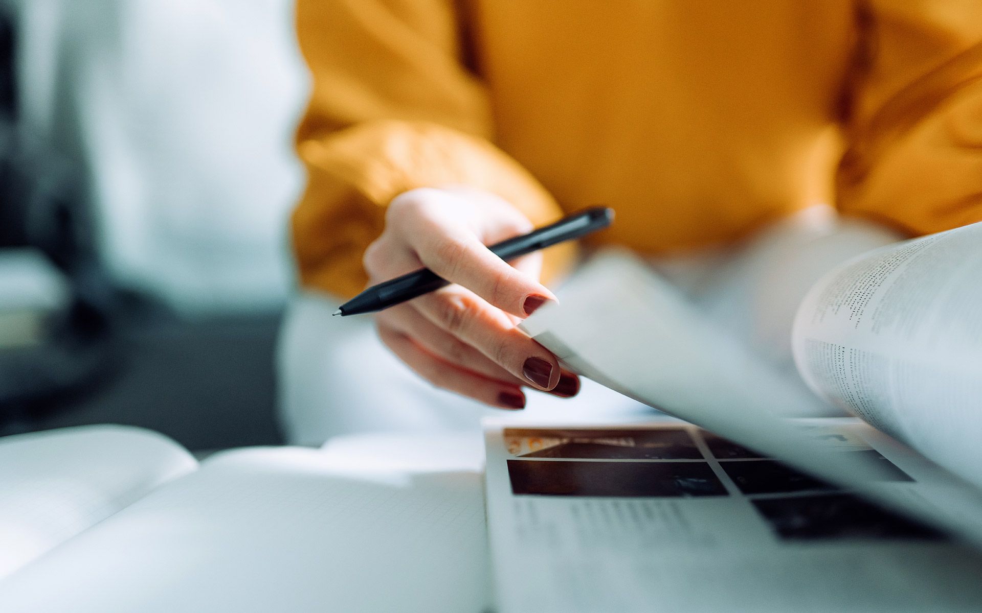 Person in yellow sweater holding pen, flipping through pages of a magazine.