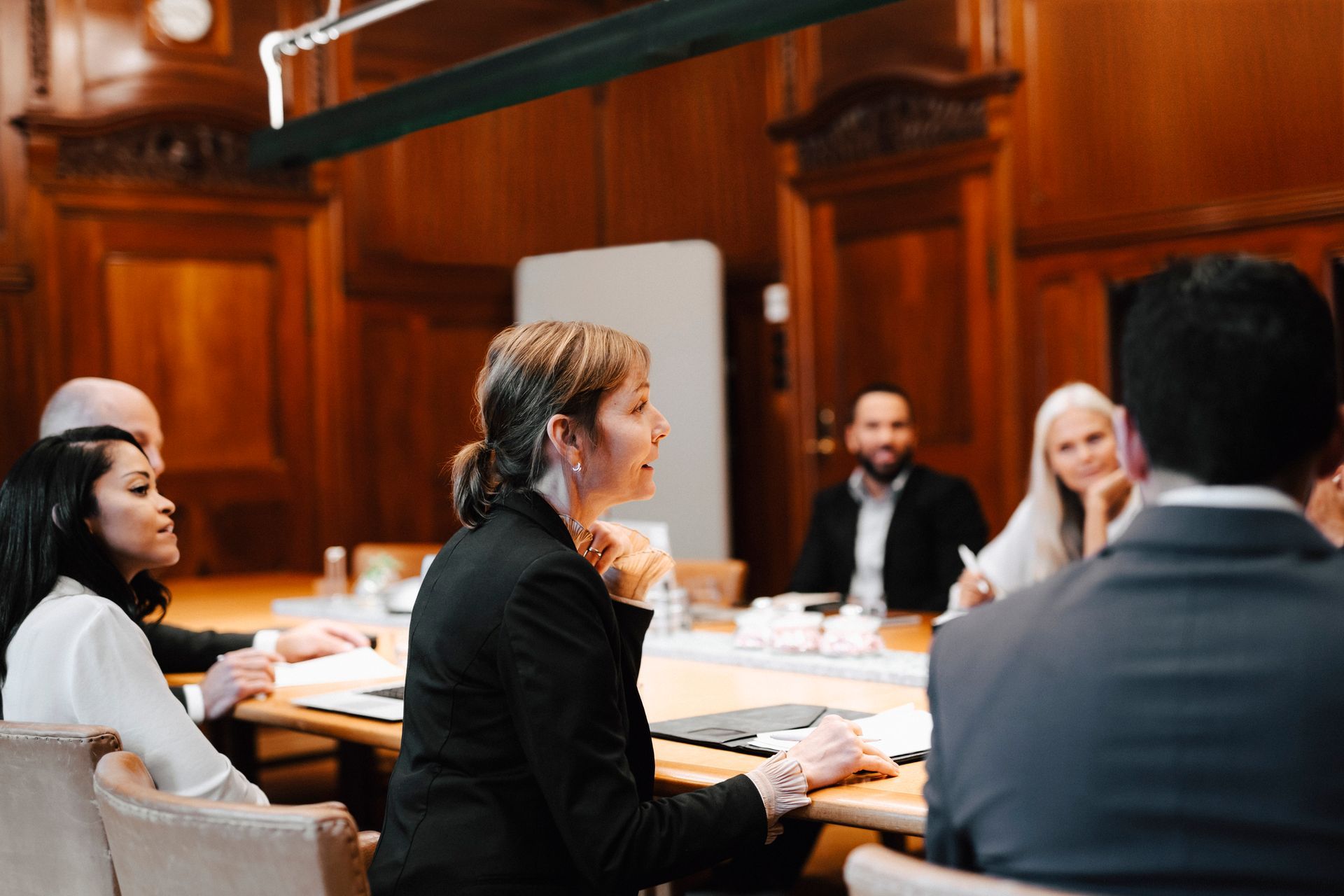 A woman speaks at a conference table, facing other attendees in a wood-paneled room.