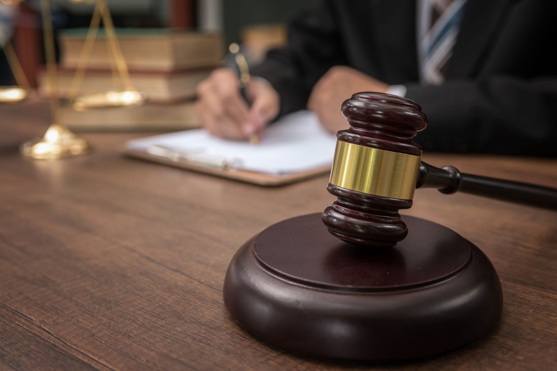 Gavel on wooden block; person in suit writing on a clipboard; scales of justice in background.