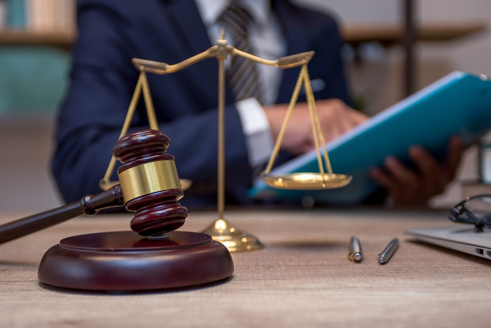 Gavel on a wooden block next to a golden scale. A person in a suit is in the background, reviewing documents.