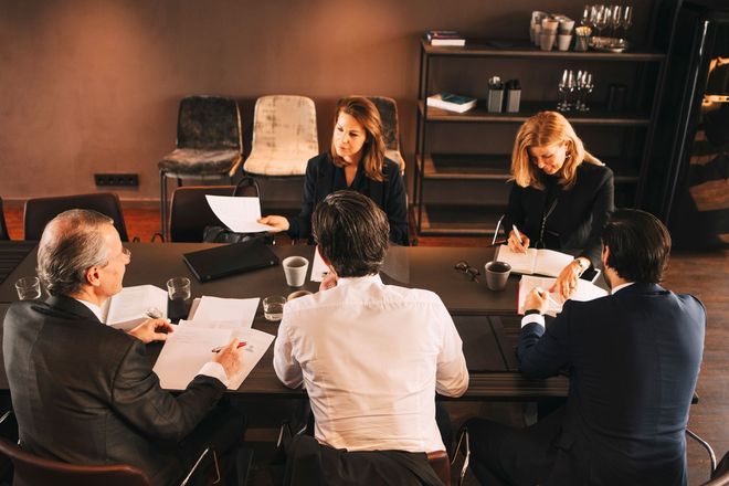 People in business attire seated around a table, reviewing documents, in a conference room.