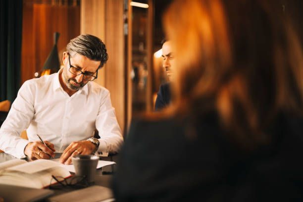 Man in white shirt signing documents at a table, two others in background.