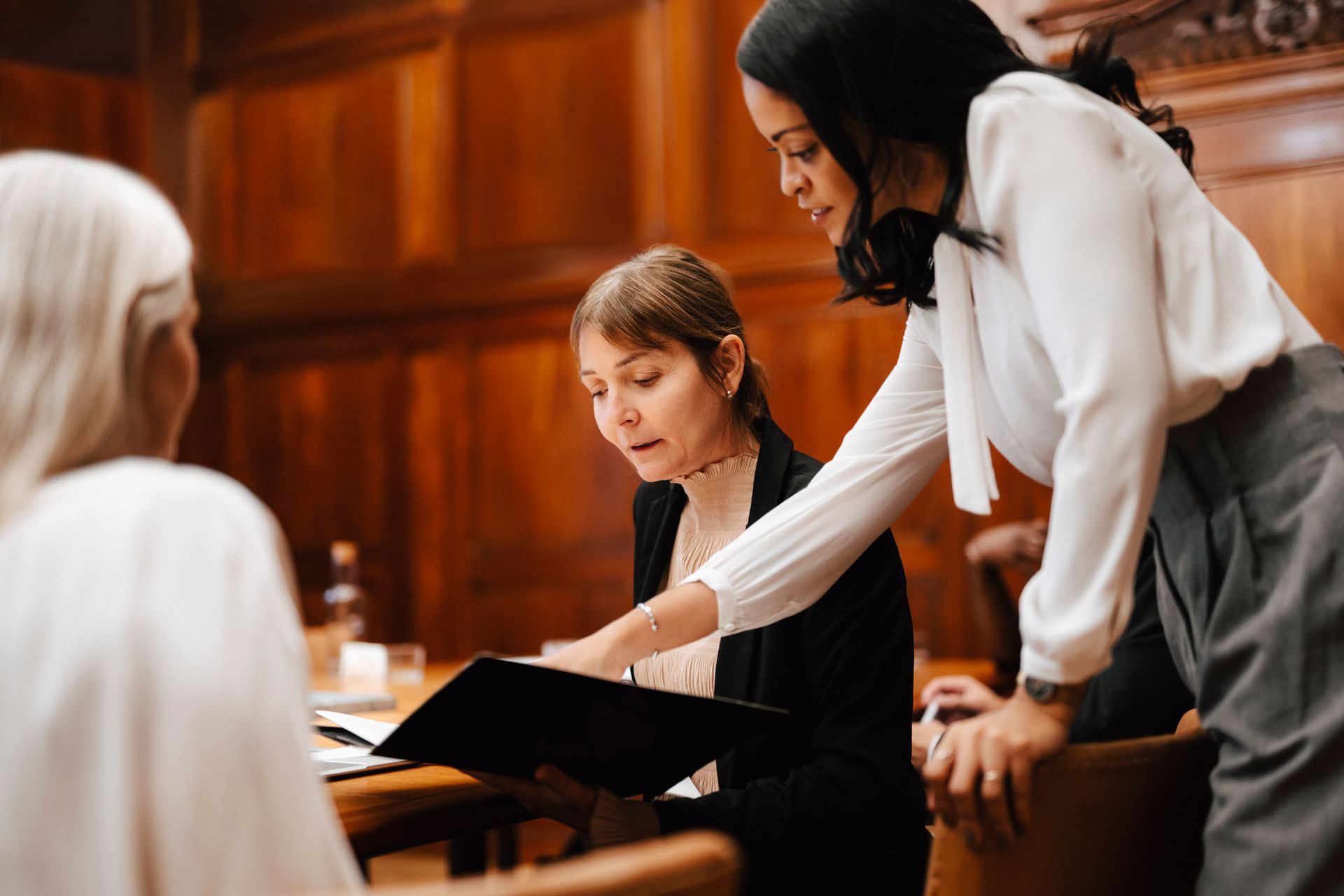 Two women review paperwork, one pointing. Another woman is seated, facing away, in a wood-paneled room.