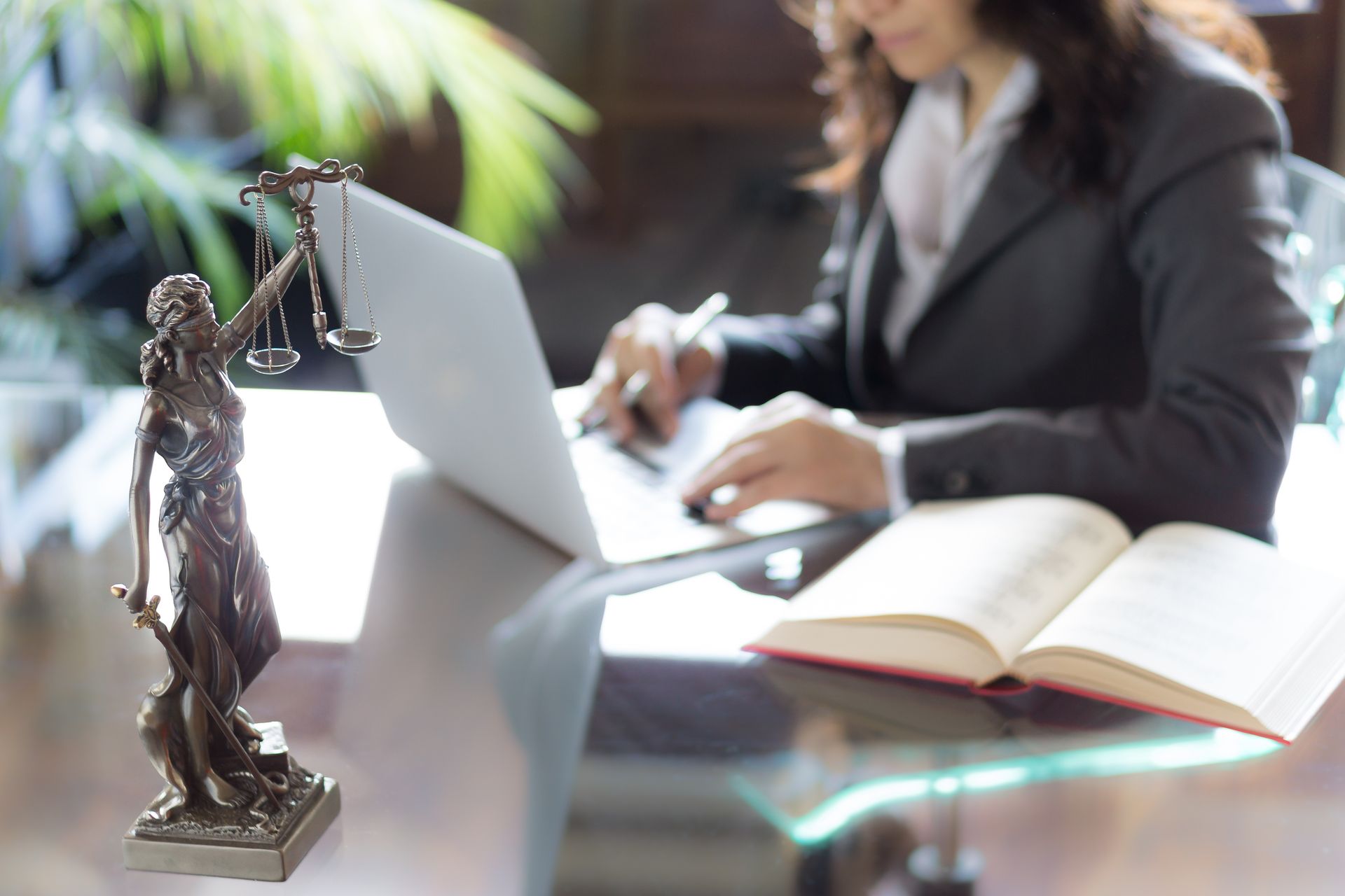 Statue of Lady Justice on desk near a person working on a laptop, with an open book.