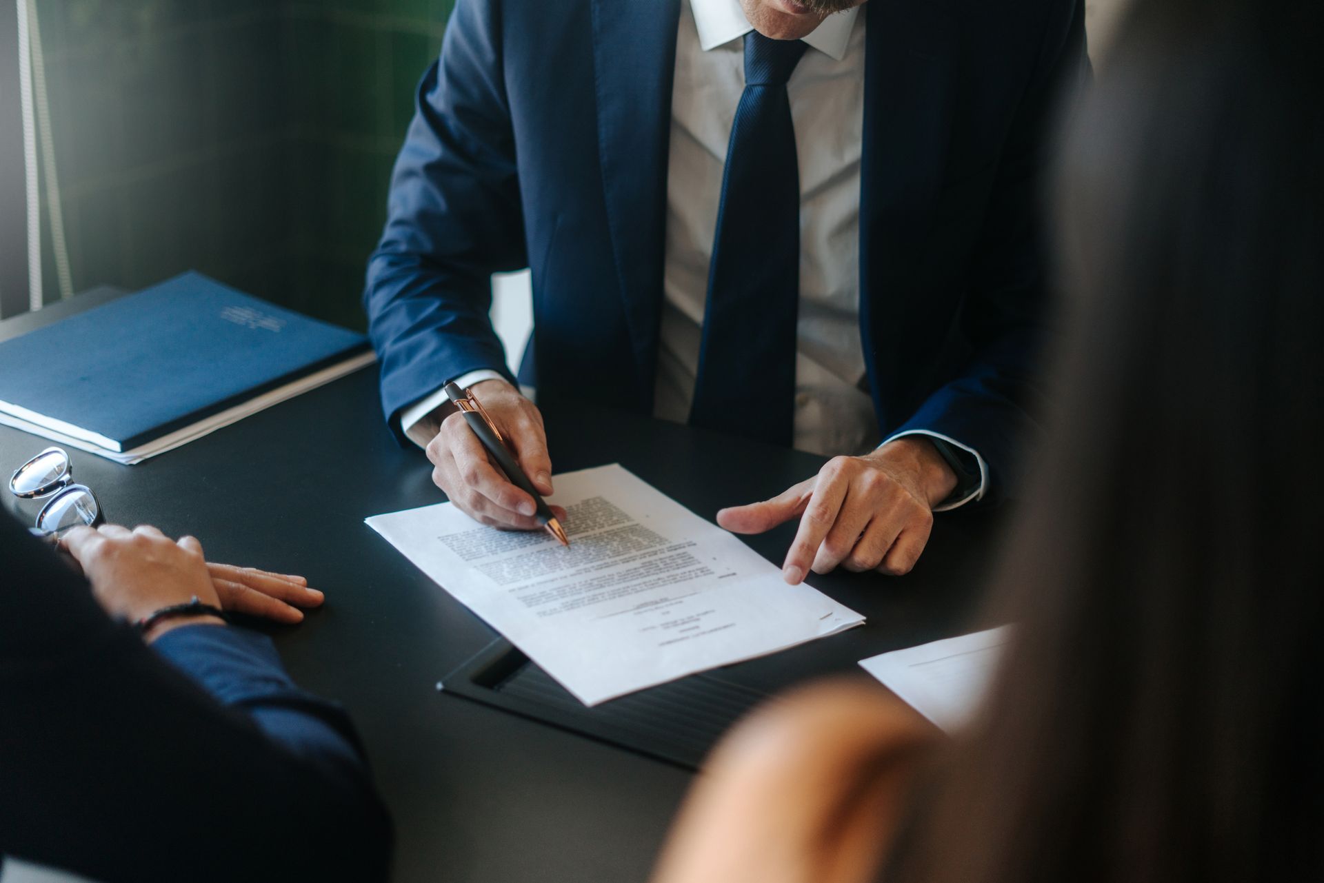 Person in suit signing a document at a table, two others observing.