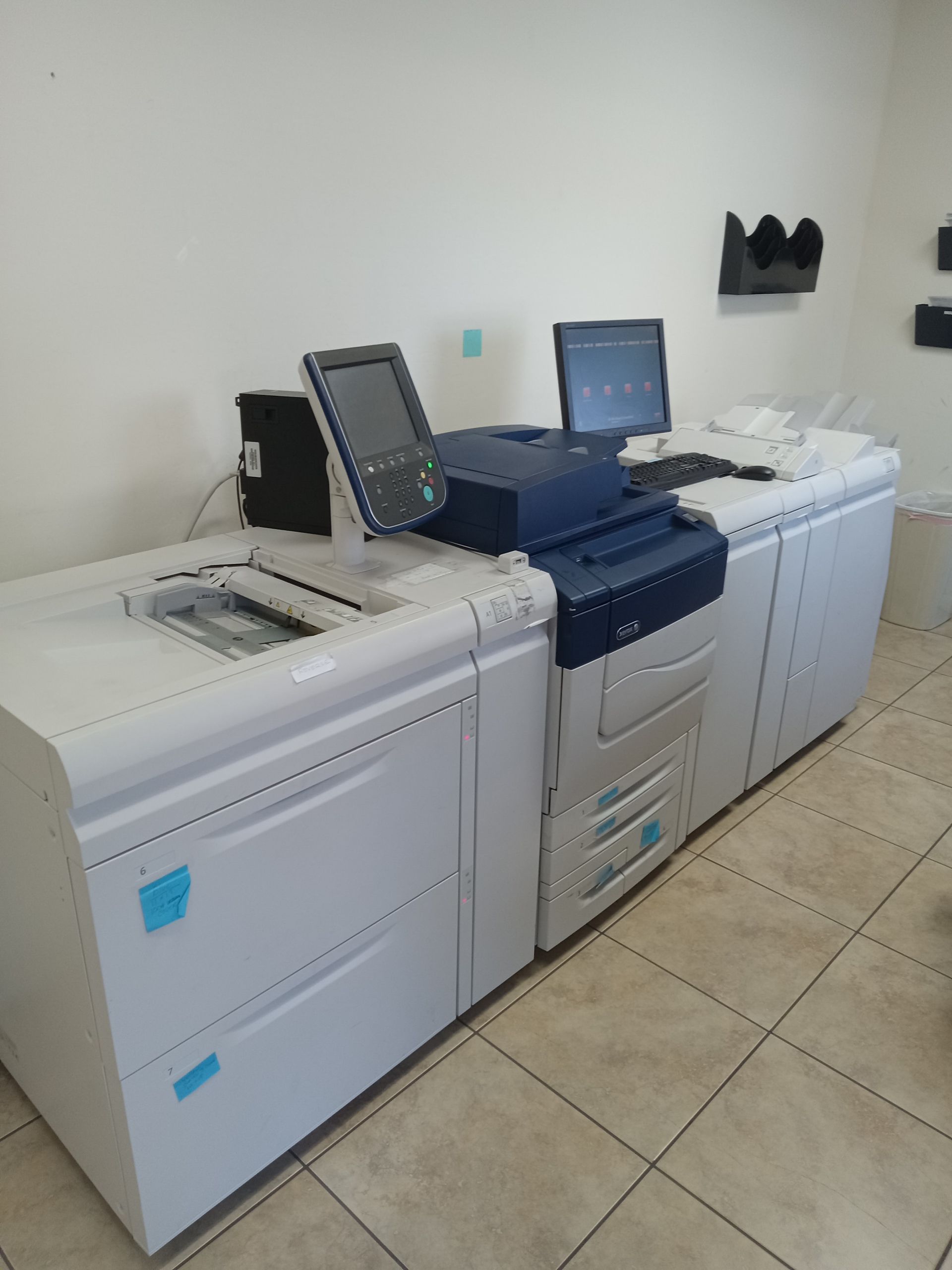 A large Xerox digital printer in a room, with control screens and paper trays, set on a tiled floor.