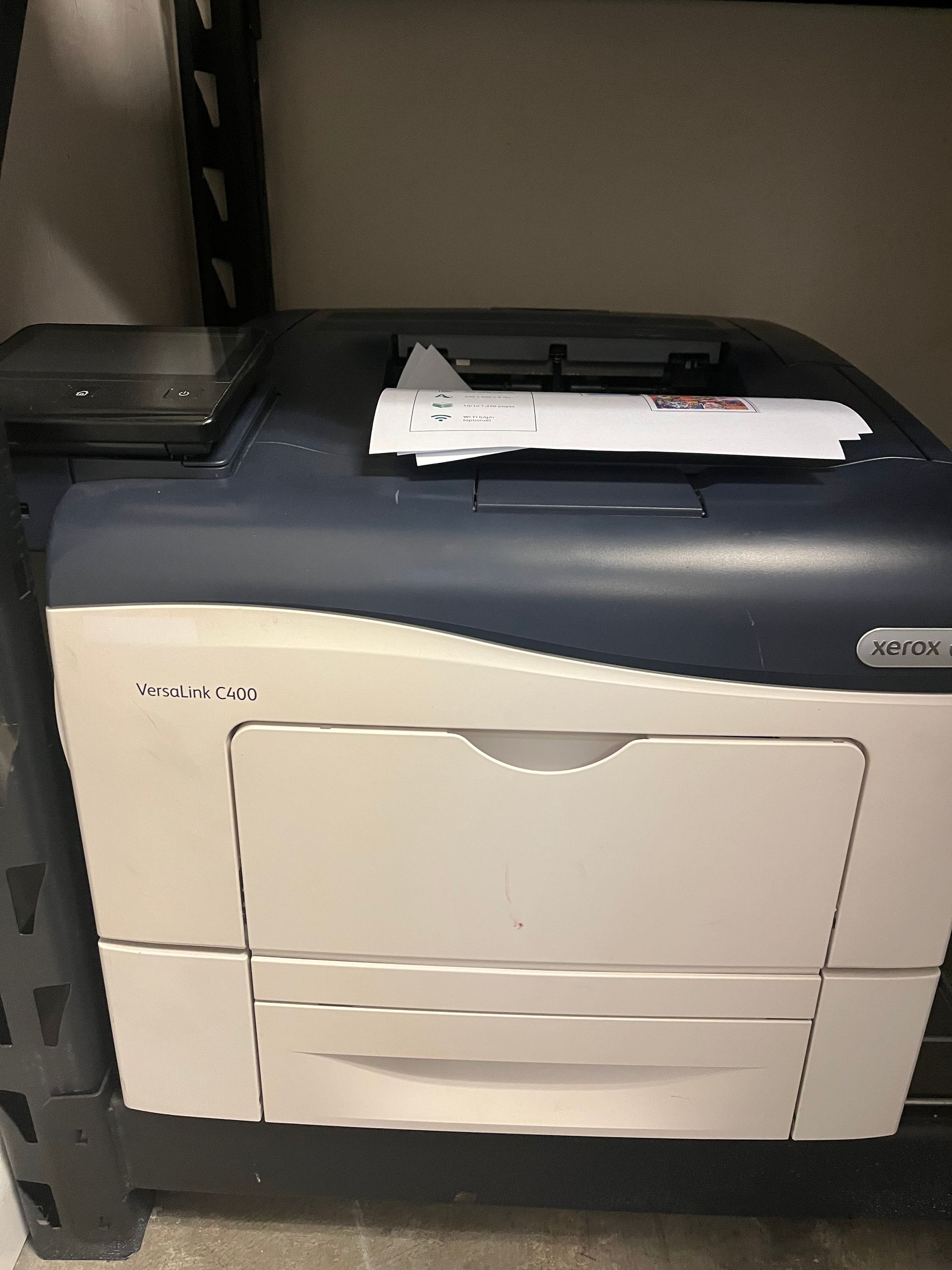 A Xerox printer on a shelf, with a stack of paper on top. It is white and navy blue.