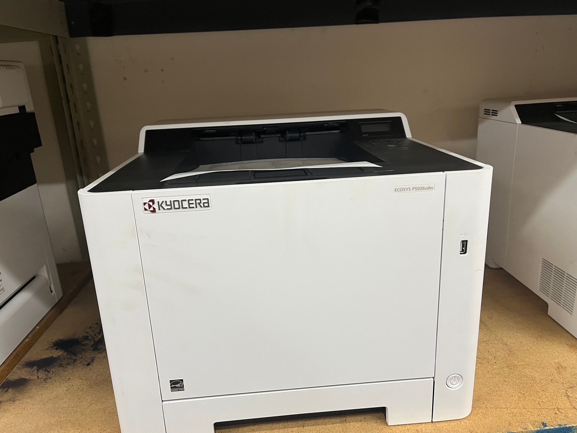 White Kyocera printer on a beige floor, with black top, in an office setting.