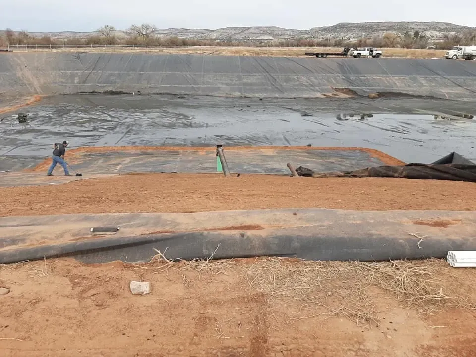 Man working in a large, tiered construction site. Brown dirt, black lining, and utility vehicles are visible.