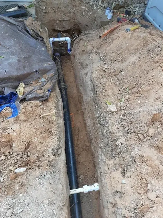 Trench with black pipe running through it, white PVC pipe connections, and exposed dirt.