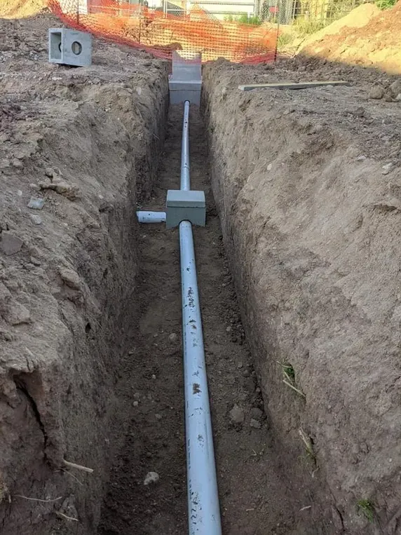 Trench with gray pipes, fittings, and concrete structures, likely for underground utility installation in an earthen area.