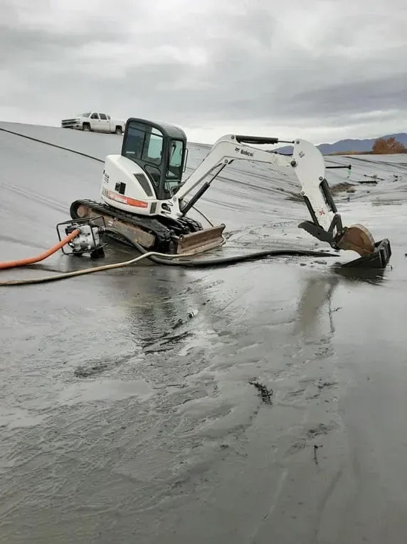 Bobcat excavator on a wet, gray surface, near a white truck, under a cloudy sky.