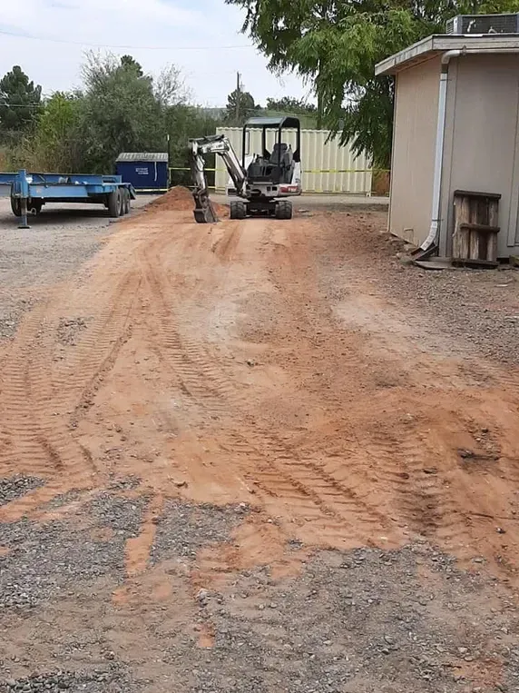 An excavator works on a dirt road next to a white building and blue trailer.