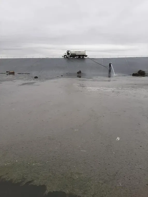 Truck driving across a body of water, while a stream of water shoots upward. Overcast sky.