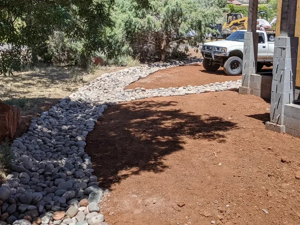 Dirt yard with rock borders, truck, trees, and construction equipment in the background.