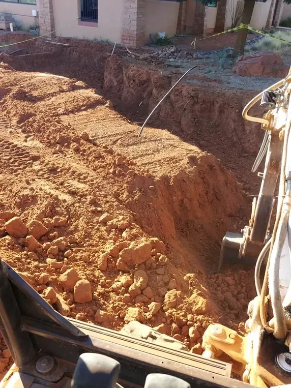 Excavator digging a trench in reddish-brown soil near a house.