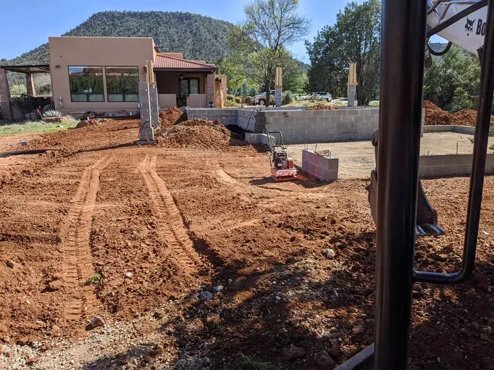 Construction site: Red dirt, foundation, small excavator, house with large windows, mountain backdrop.