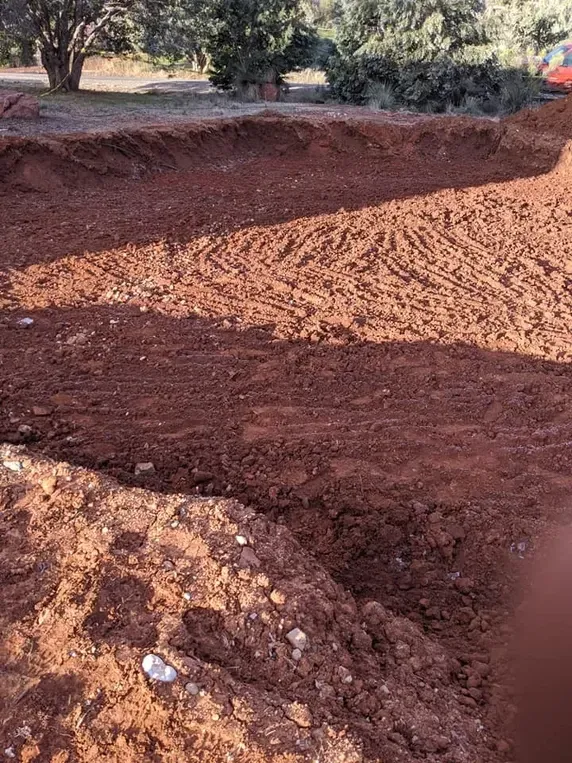 A dug-out pit with red-brown dirt, potentially for construction, under sunny conditions.