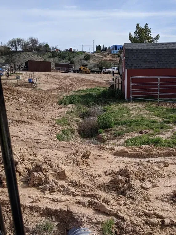 Muddy lot with red barn, construction equipment, and a blue building in the background.