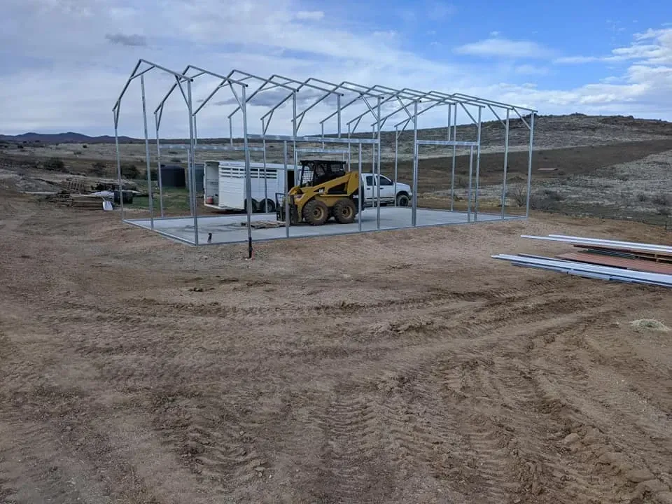 Metal frame of a building under construction on a concrete pad. A skid steer and truck are present.