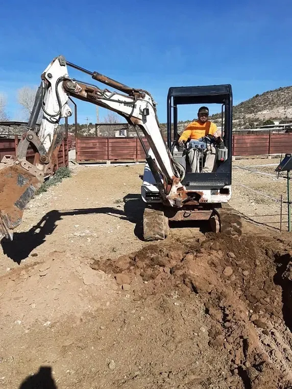 A person operates a white excavator, digging dirt. They are wearing an orange jacket on a sunny day.