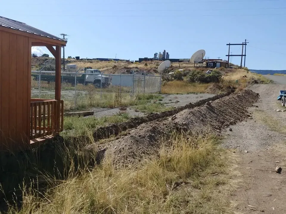 A trench dug near a wooden building, a chain-link fence, and satellite dishes under a blue sky.