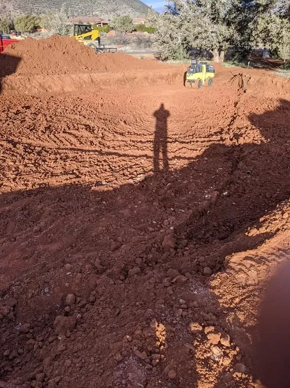 Construction site with red soil, excavators, and shadow of person.