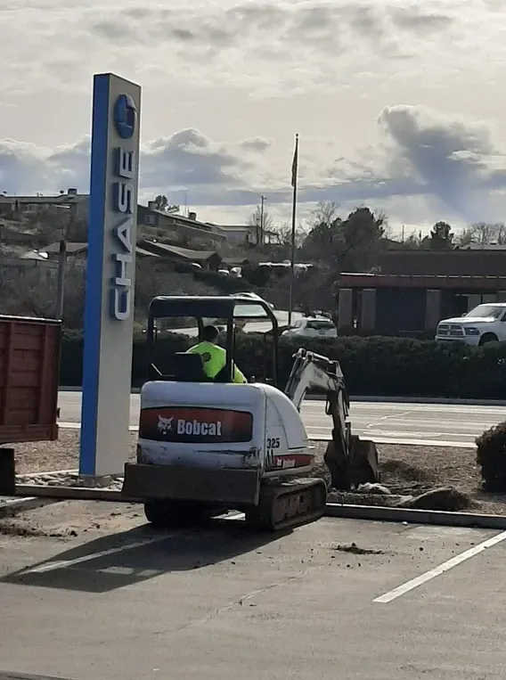 A Bobcat excavator digs near a Chase bank sign. A worker in a green vest operates the machine.